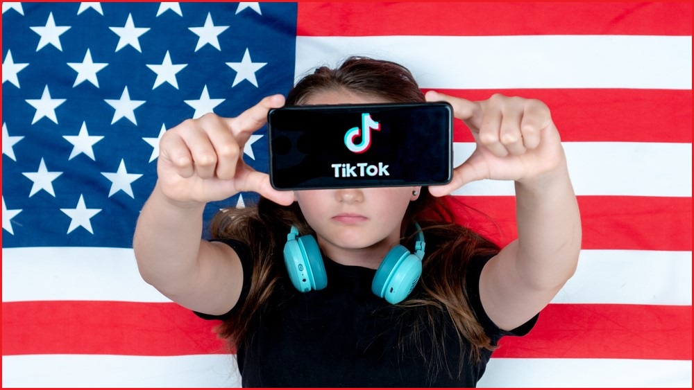 Young person standing in front of US flag holding phone displaying TikTok logo.
