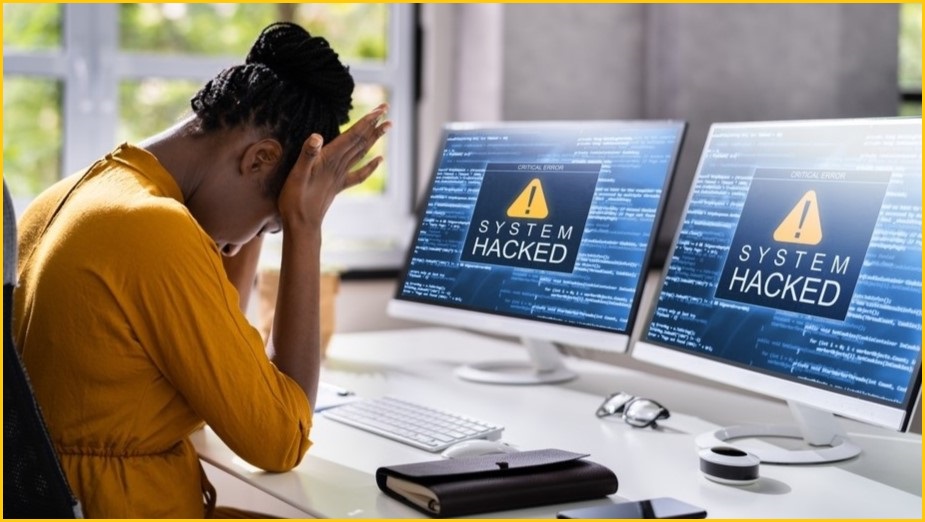woman holding her head in her hands after the computer screen in front of her show SYSTEM HACKED message