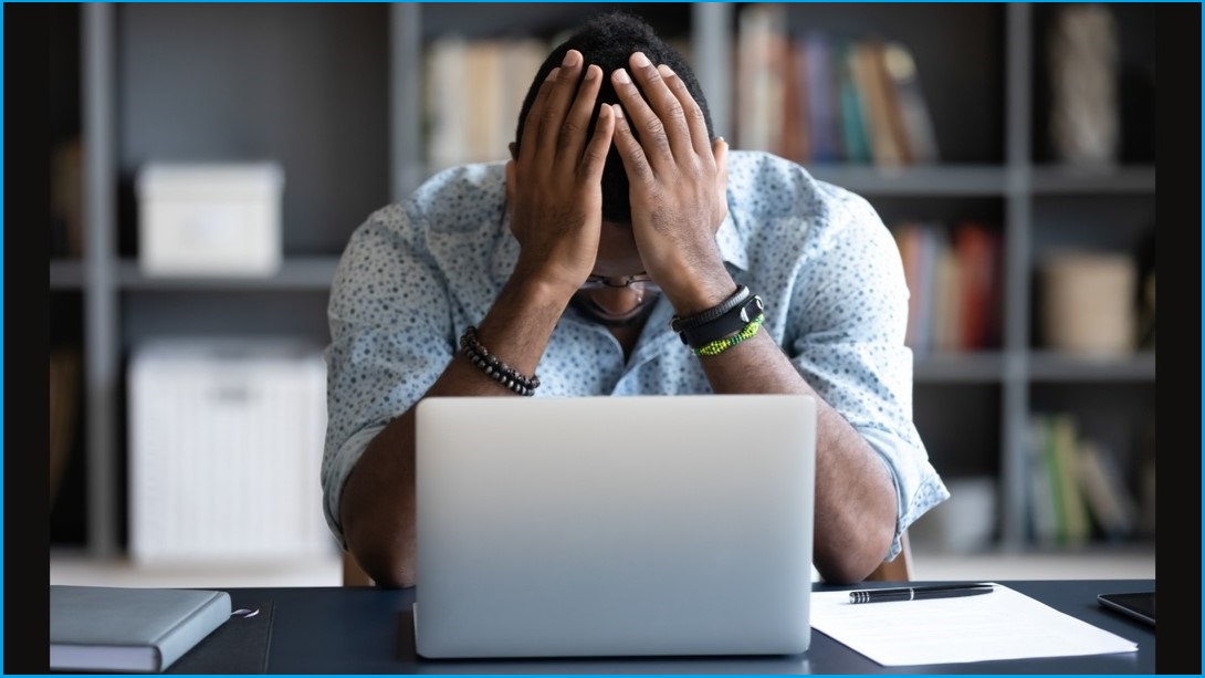 Distraught man sitting face a computer screen, holding his face in his hands.