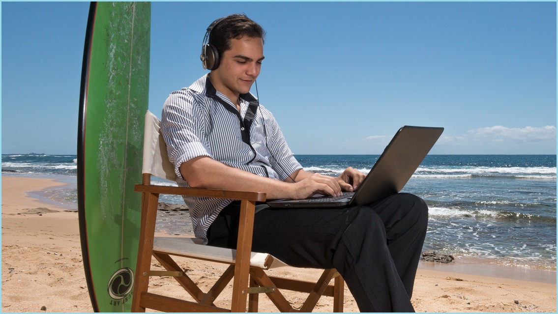 Man working on a laptop, sitting on a beach with a surfboard behind him
