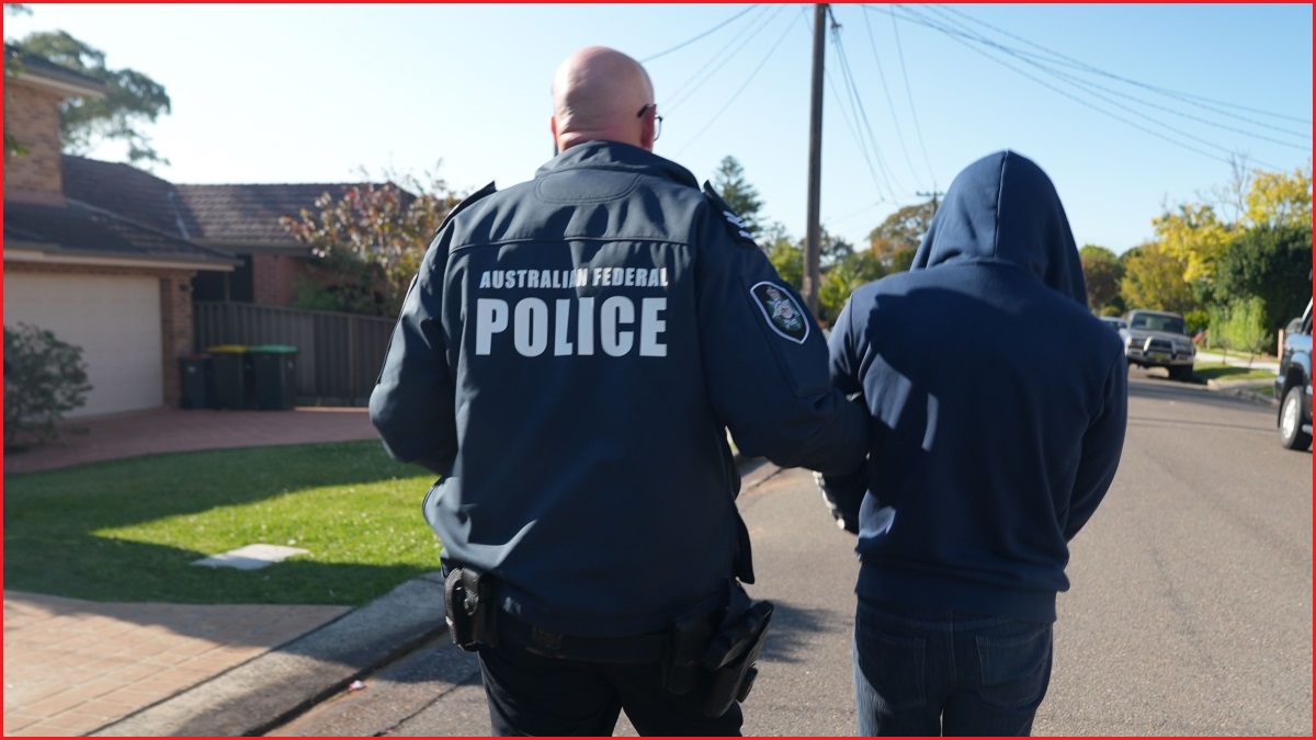 A medium shot behind a policeman holding a man by the arm and walking him down a suburban street.