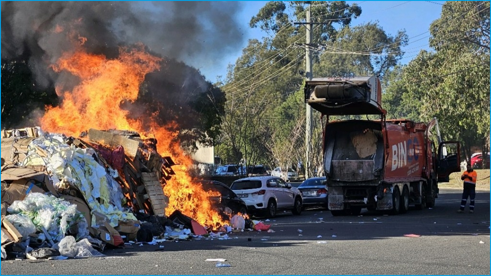 A wide shot of a garbage truck stopped on a street next to a large pile of garbage which is on fire.