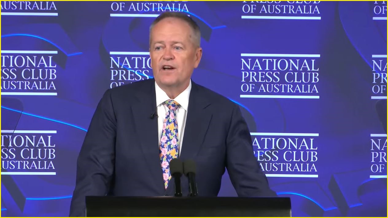 A medium shot of Bill Shorten, middle-aged man in a suit and tie speaking at a podium into a microphone.