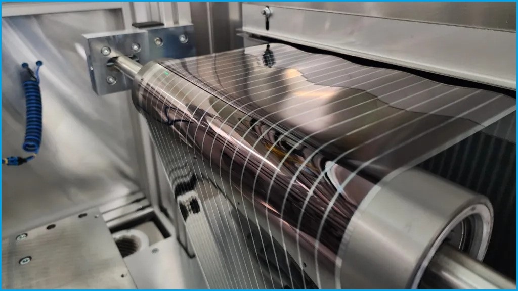 A close up of a roll of flexible solar panel moving through a metal machine in a factory.