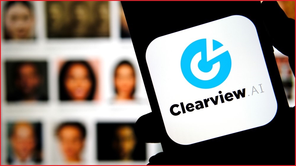 A close up of a mobile phone displaying the Clearview AI logo. Behind, a backdrop of images showing various people's faces.