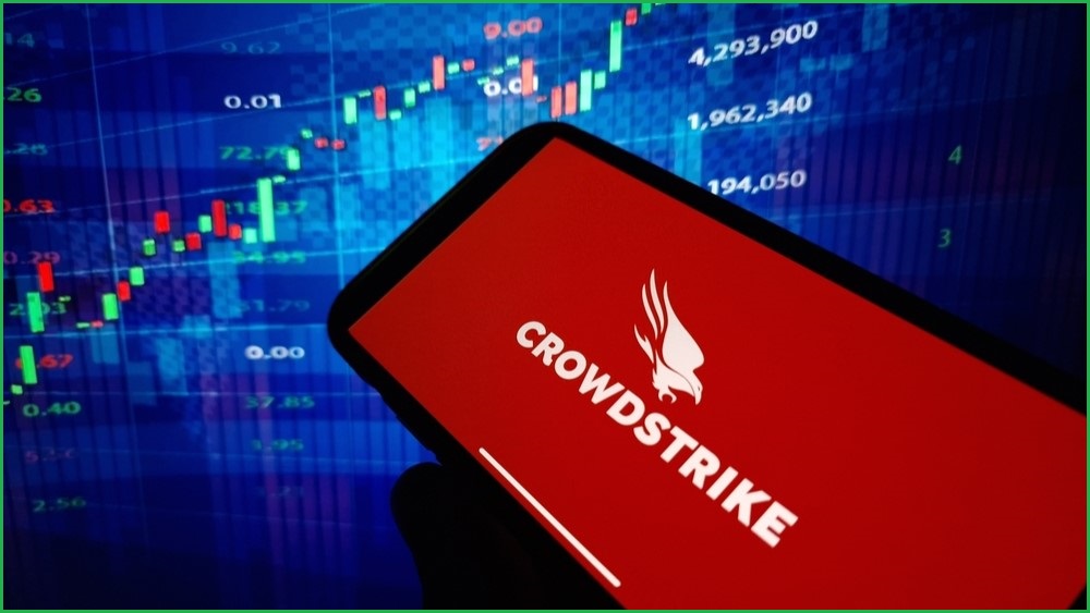 A close up of a smartphone displaying the CrowdStrike logo on its screen. In the background, a large screen displaying a share price chart.