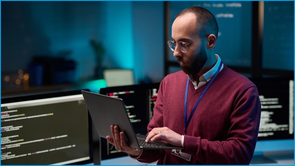 A medium shot of a balding man in glasses holding and looking at a laptop in a room filled with computer screens.