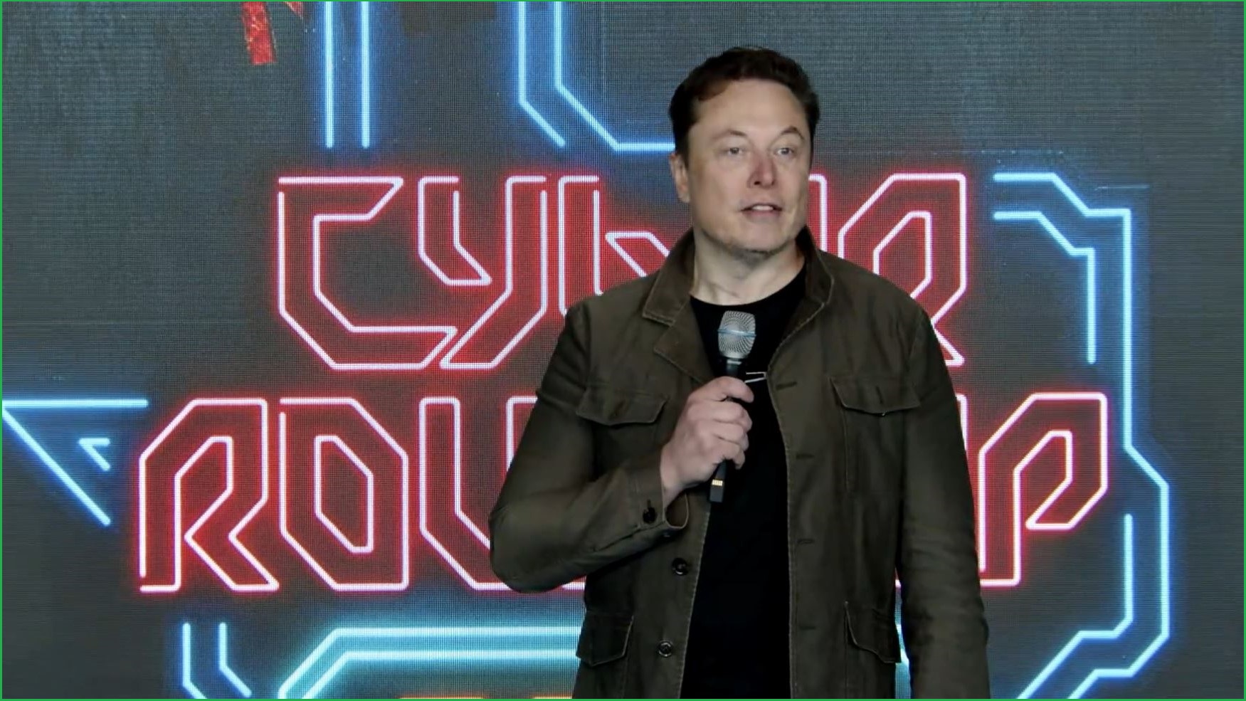 Elon Musk stands on a stage speaking into a microphone, in front of an electronic sign with the words 'Cyber roundup'.