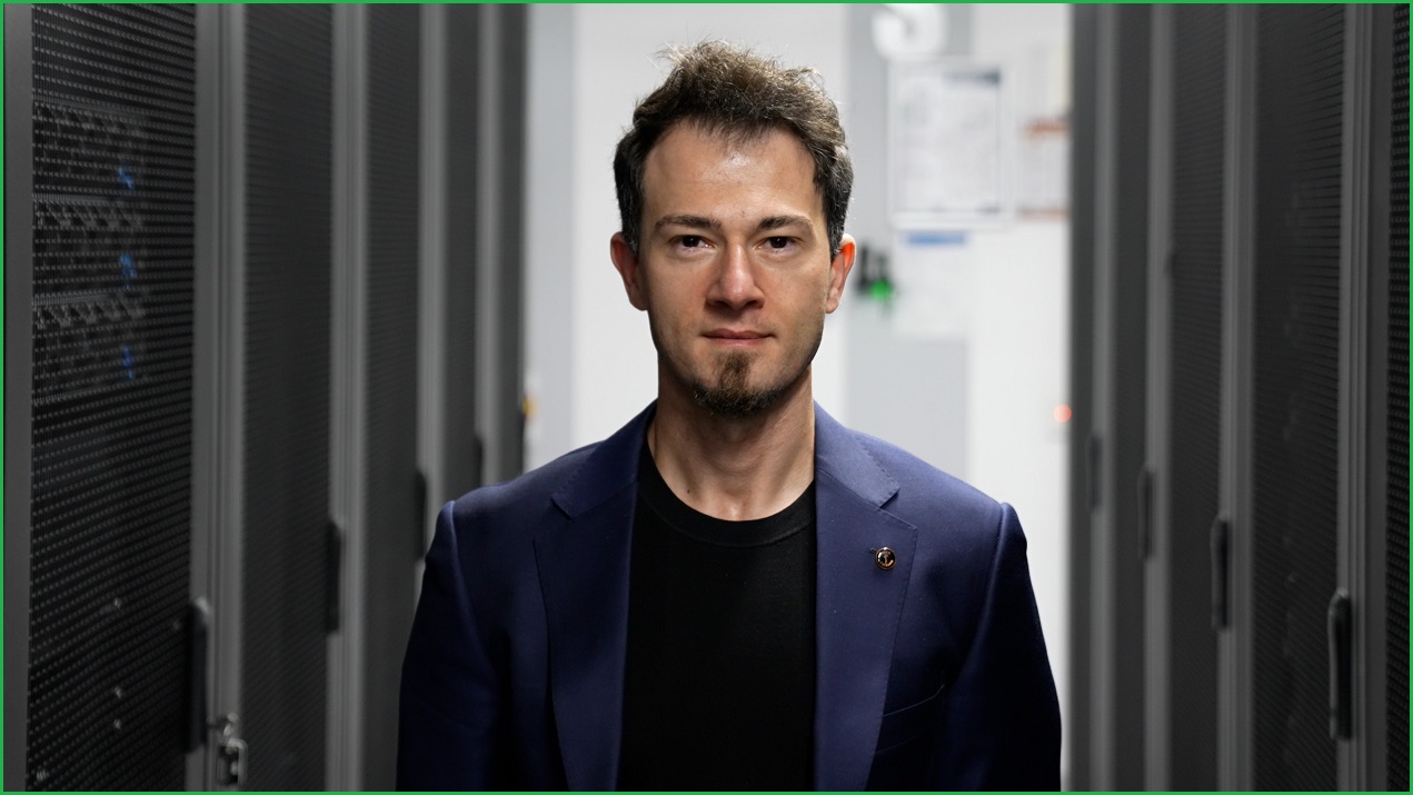 A close up of a man in a suit jacket standing between walls of computer servers.