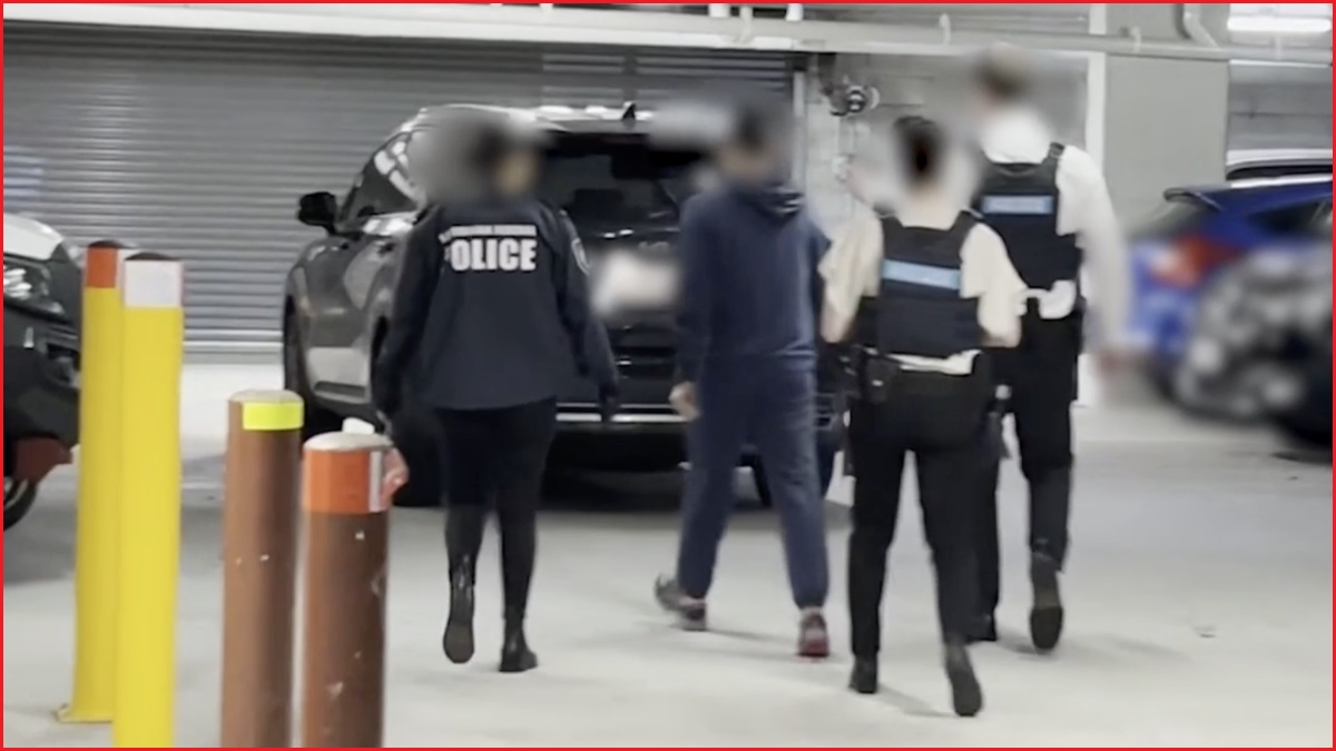 Police in uniforms and a person wearing a hoodie walk towards a car inside a car park.