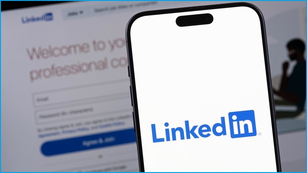 A close up of a smartphone displaying the LinkedIn logo, in front of a computer screen displaying the LinkedIn website.