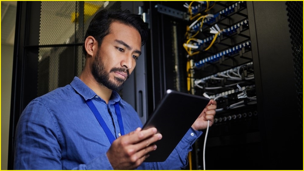 A man standing next to a server rack hold and looks at a tablet, while holding a wire connected to the server with his other hand.