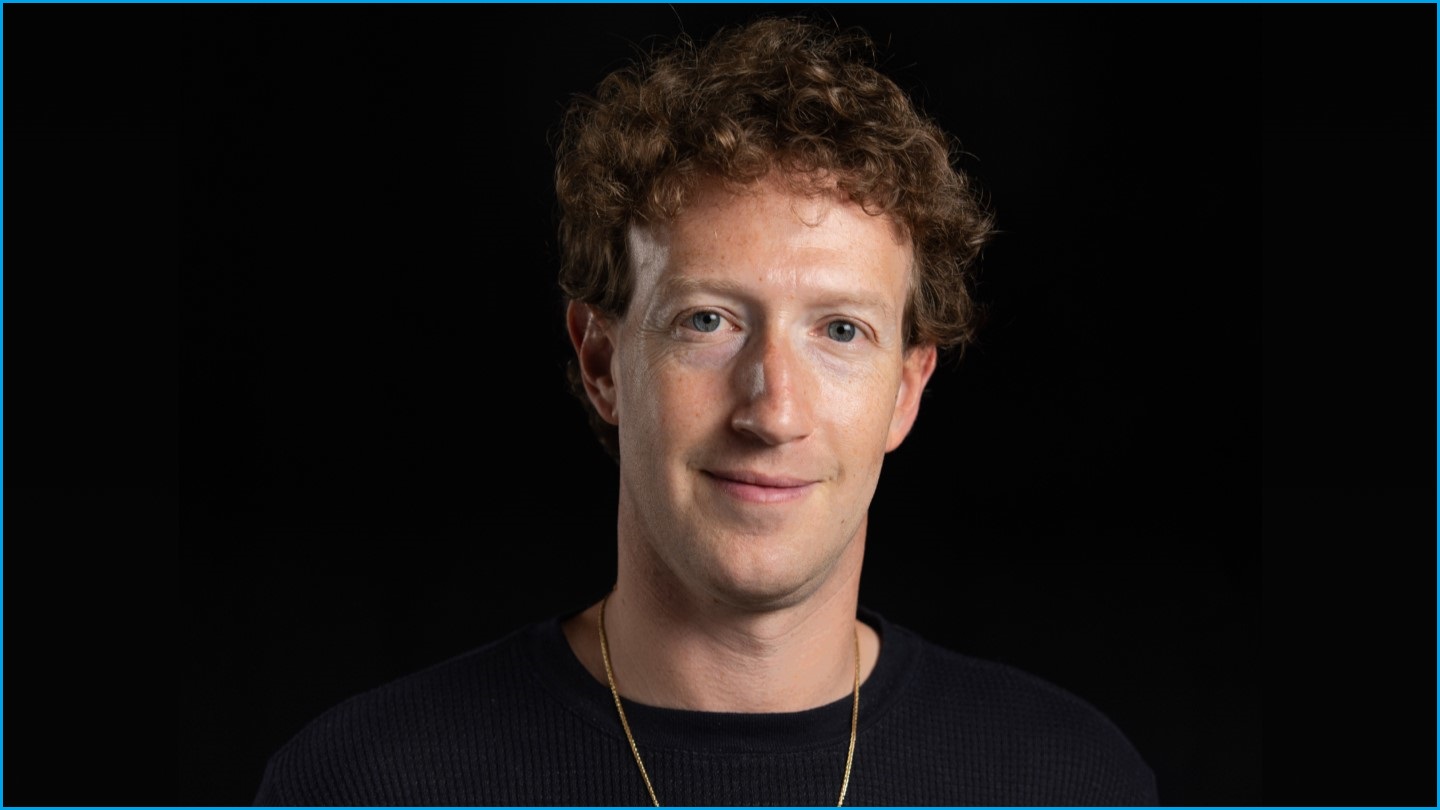 A close up of Mark Zuckerberg, smiling slightly and wearing a gold chain and dark T-shirt.