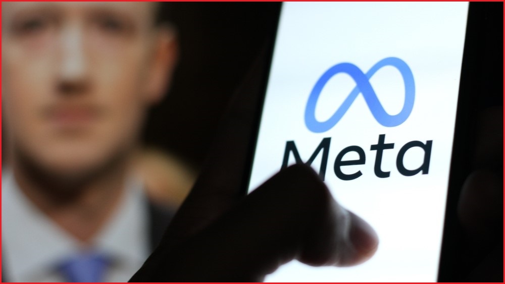 A close up of a hand using a smartphone which has the Meta logo displayed on its screen. A blurred face of Mark Zuckerberg in the background.