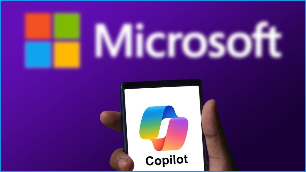 A hand holding a smartphone displaying the Microsoft Copilot logo, in front of a backdrop showing the full Microsoft logo on a wall