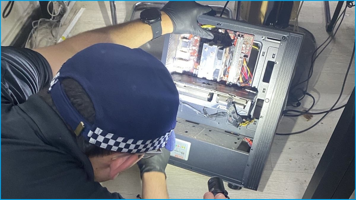 A close up of a policeman in a police hat wearing gloves and holding a torch as he looks inside a desktop computer.