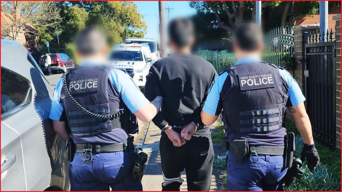 A medium shot of the backs of two police officers holding a man between them, as they walk down a footpath.