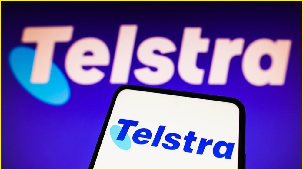 A close up of a smartphone displaying the Telstra logo, in front of a backdrop of a larger Telstra logo.