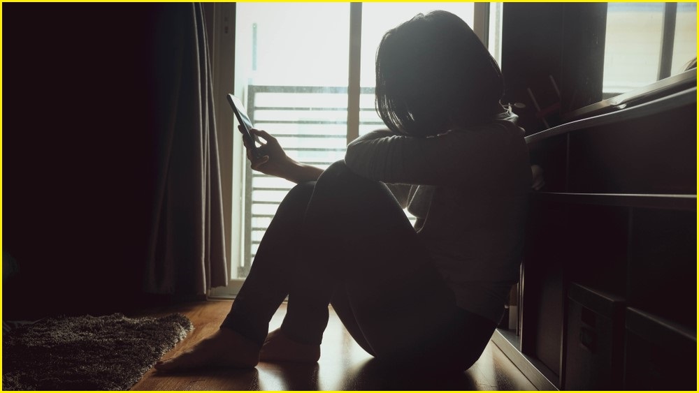 A silhouette of a woman sitting on a wooden floor, looking at her mobile phone in the dark.