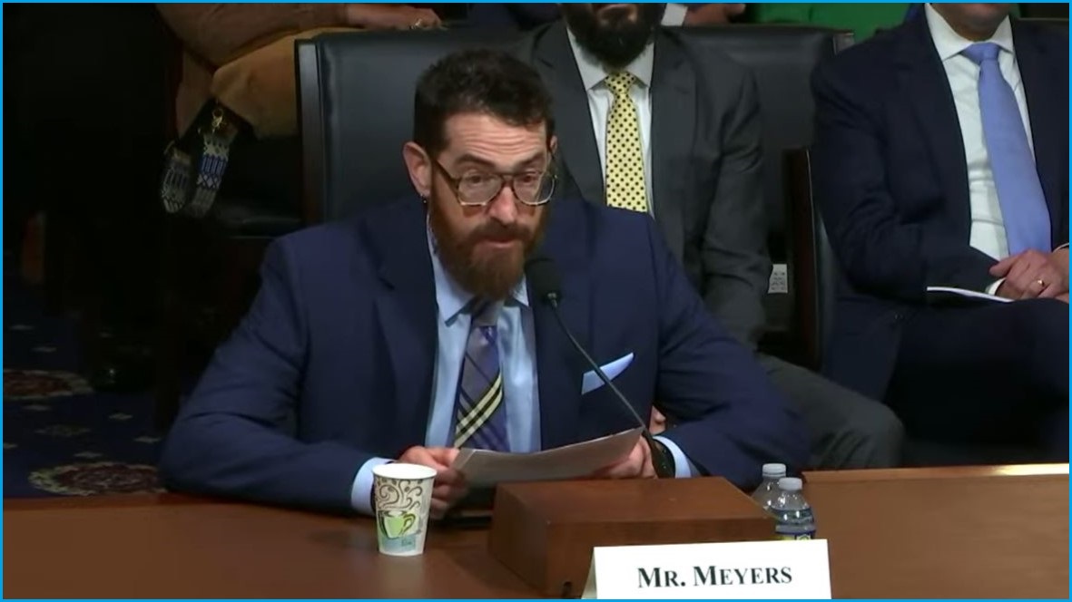 A medium shot of a man with a beard and glasses sitting at a table holding paper and speaking into a microphone.