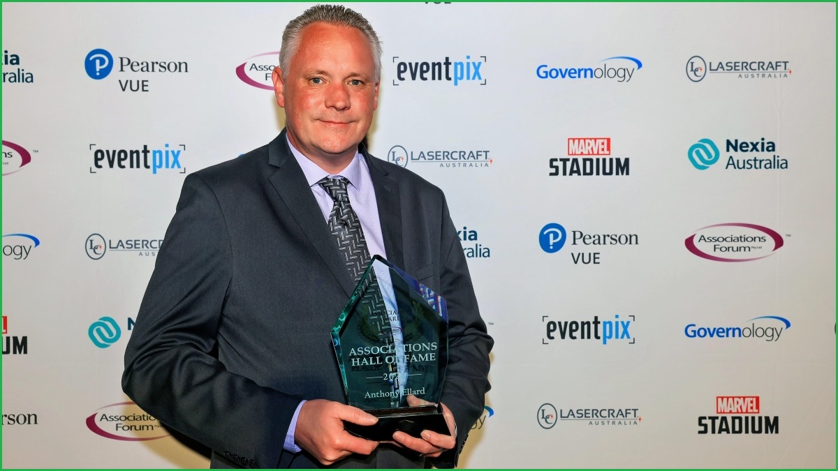 A medium shot of a middle-aged man in a suit holding an award and smiling, in front of a photo wall of sponsor logos.