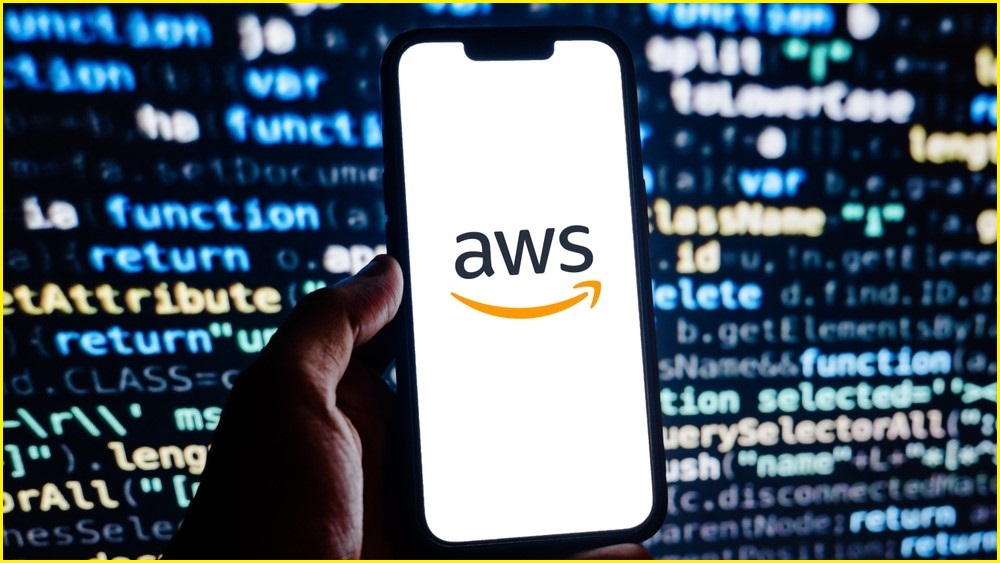 A close up of a hand holding a smartphone with the AWS logo on it. Computer code in the background.