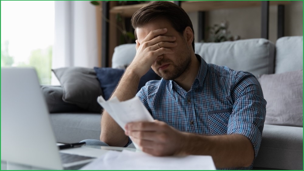 Man sitting on front of a computer, with one hand holding a bill and the other covering his eyes.