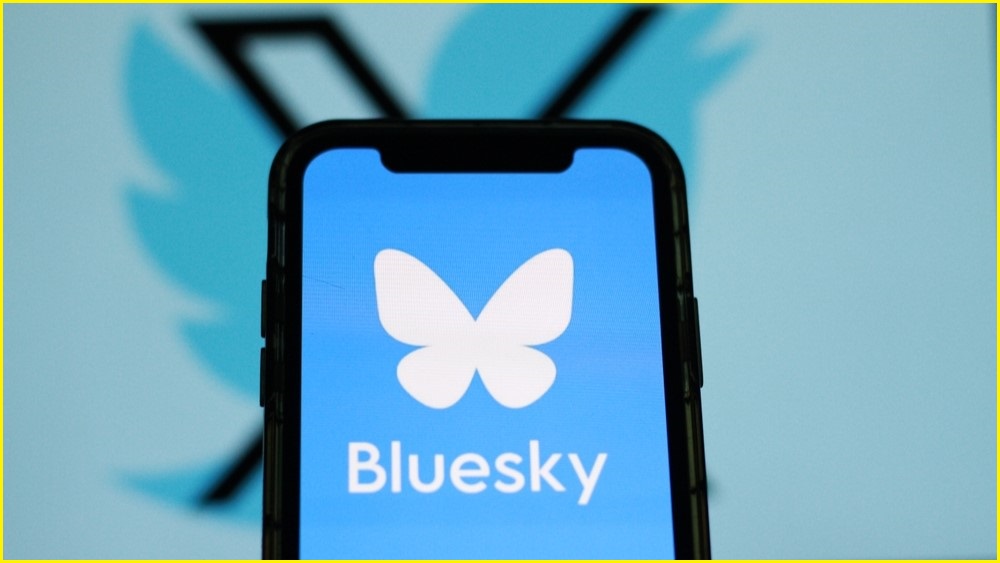 A close up of a smartphone displaying the Bluesky logo, with the X and Twitter logos on a screen in the far background.