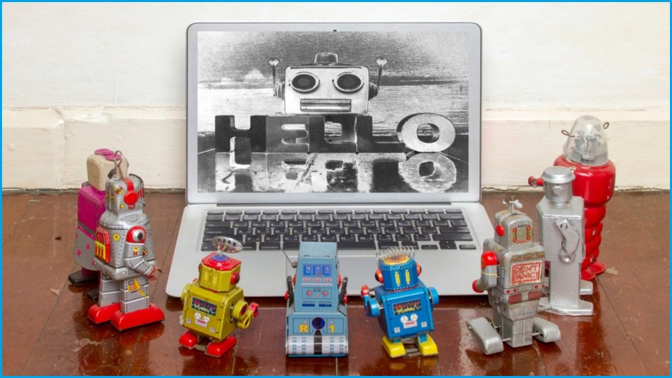 A bunch of small robots staring at a large robot on a laptop screen.