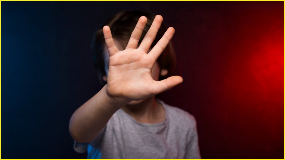 A close up of a young boy's hand, which he is holding up to cover his face as he stands in a dark space.