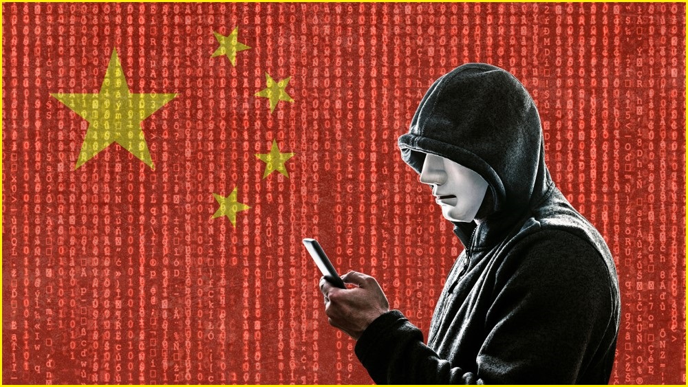 A medium shot of a male in a hoodie holding a phone and wearing a mask, standing in front of a large Chinese flag.