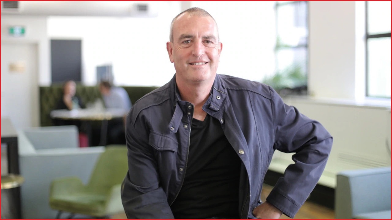 A medium shot of a bald middle-aged man smiling, with one hand on his hip, inside an office with people sitting in the background.