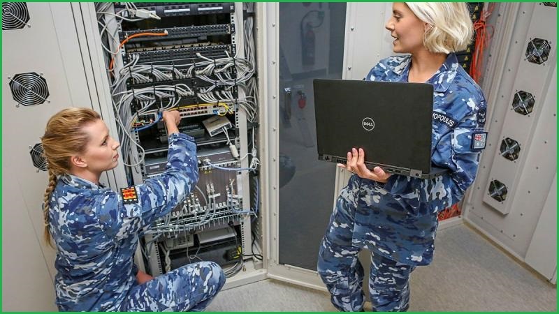 Two women in military uniforms crouch and stand next to a computer server, the one standing is holding a laptop.