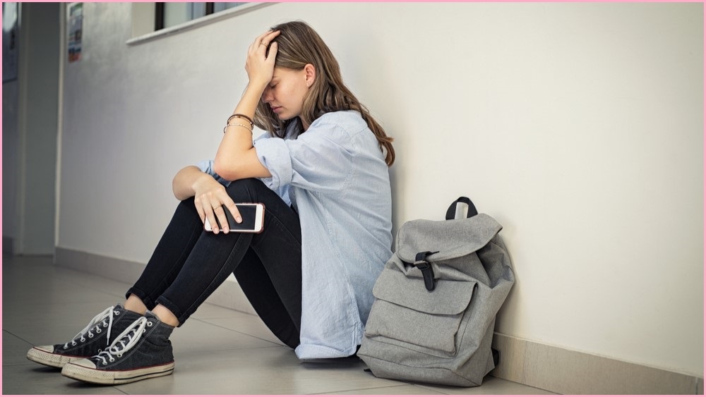 Despairing girl sitting against a wall, holding her phone and with her backpack nearby