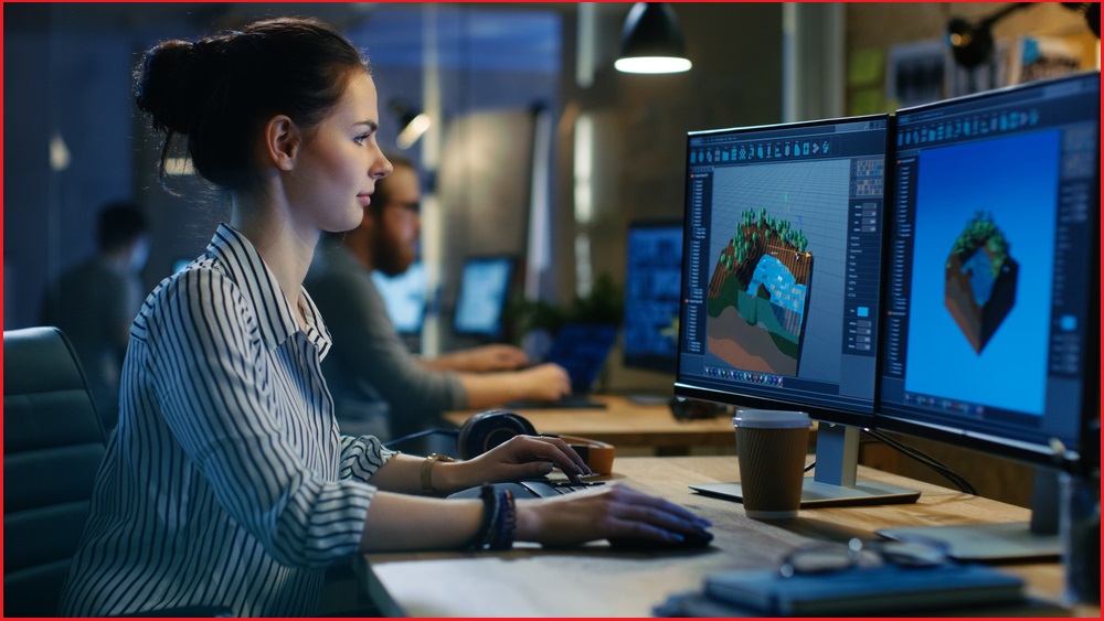 A female game developer sits at a desk and uses two computer screens to work on a computer game level.