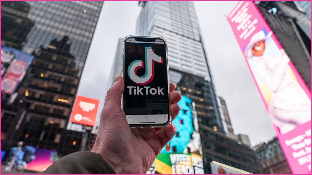 Person holding a phone up with the TikTok logo on the screen, set on a backdrop of tall city buildings.