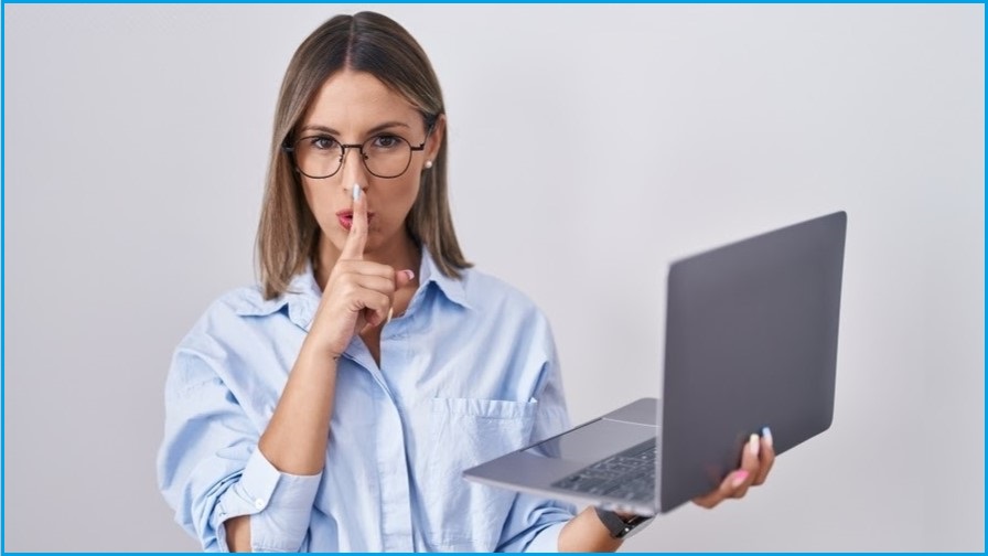 Woman in blue shirt holding laptop and a finger up to her hushed lips.