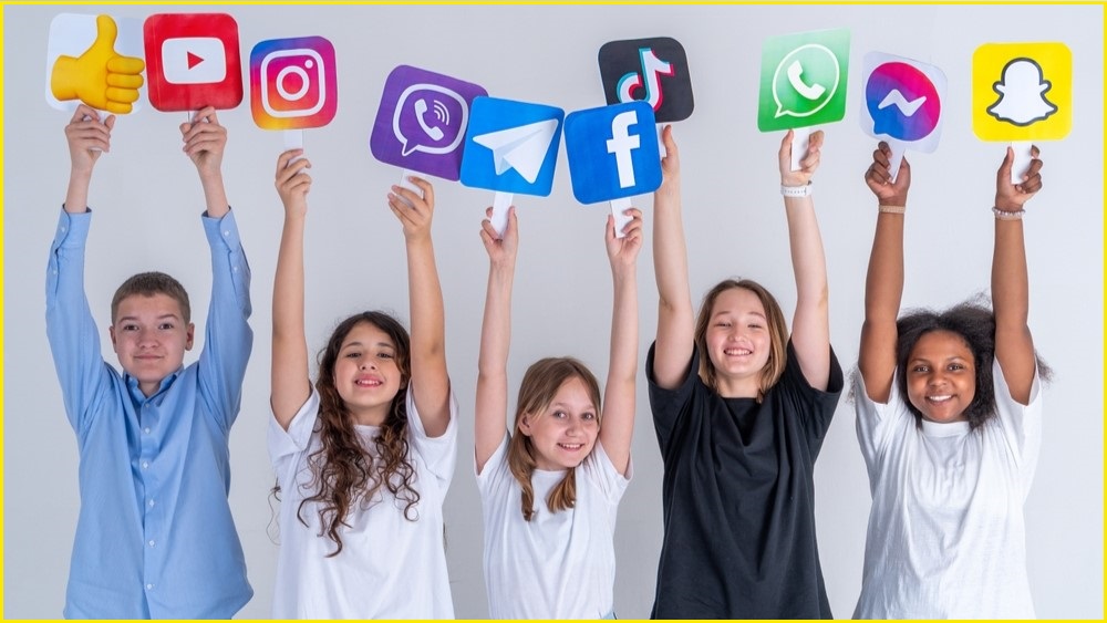 A medium shot of a group of five teenagers smiling and holding up logos of various social media platforms.