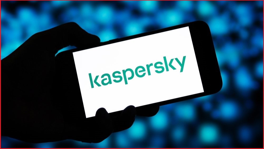 A close up of a silhoutte of a hand holding a smartphone, on which the screen displays the Kaspersky logo.
