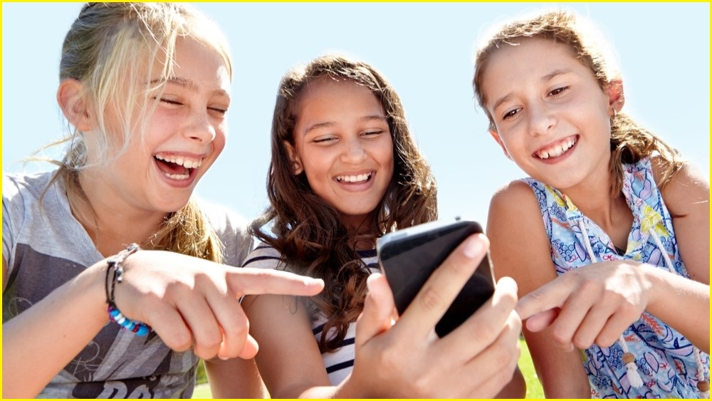 Three girls looking at a phone screen, pointing and laughing.