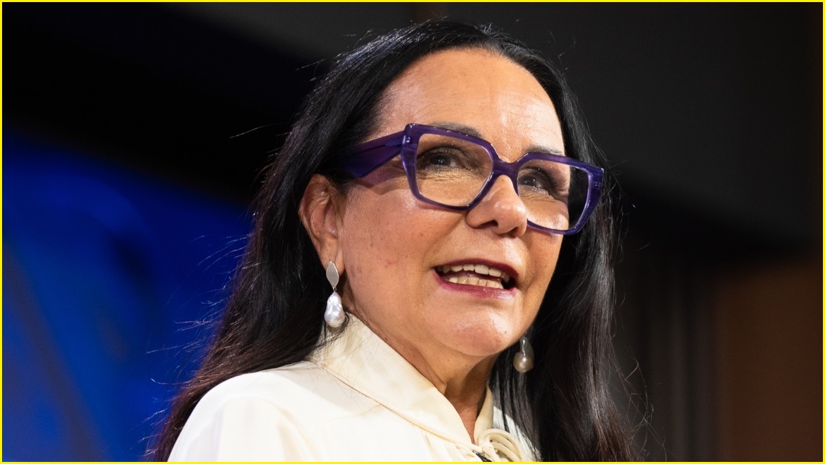 A close up of a woman in her 60s with long, dark hair, earrings and thick-rimmed glasses, speaking.