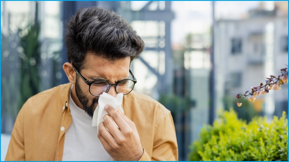 A medium shot of a man wearing glasses and walking outside blowing his nose with a tissue.