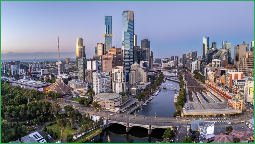 A wide shot of Melbourne's skyline at dusk, with a park and bridge over a river in the foreground, and buildings in the background.