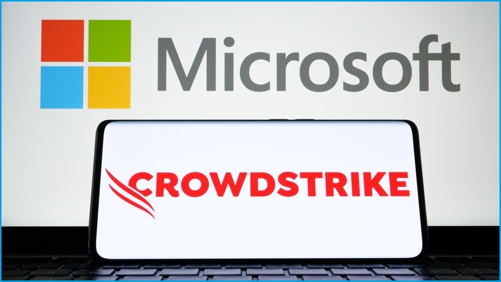 A mobile phone on its side displaying the CrowdStrike logo, laying against a laptop screen displaying the Microsoft logo.