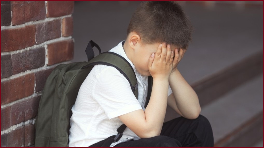 Young boy wearing a backpack leaning against a brick wall, covering his eyes.