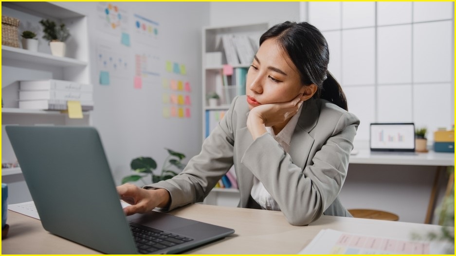 A medium shot of a woman of asian descene using a laptop at a desk while holding on hand to her face and looking tired.