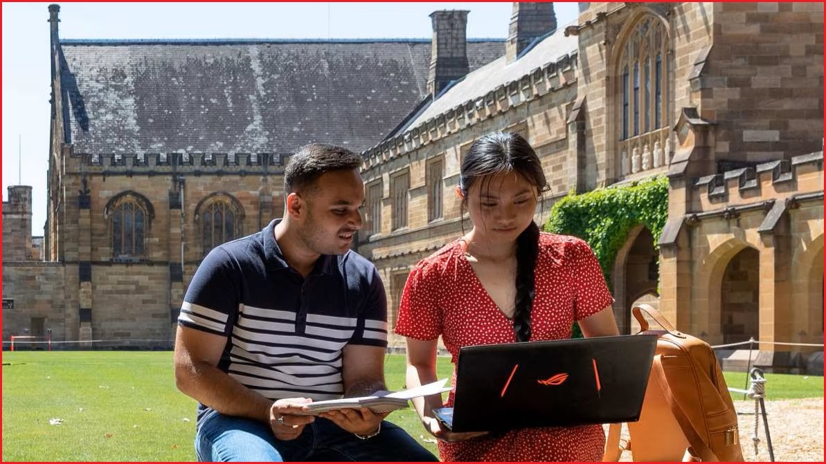 A medium outdoor photo of a young man and woman sitting on a short wall and looking at a laptop inside the University of Sydney, with sandstone buildings in the background.