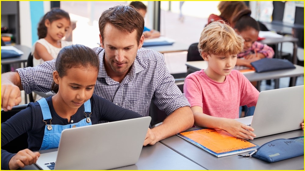 A medium shot of a male teacher crouching and helping a young girl use a laptop during class. A boy beside them also uses a laptop.