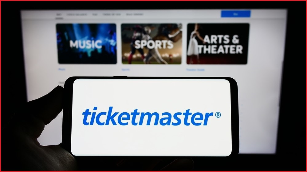 A close up of a hand holding a smartphone with the Ticketmaster logo on it, in front of a computer screen showing the Ticketmaster website.
