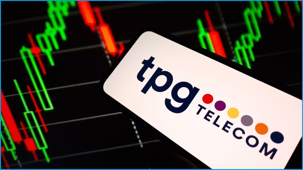 A close up of a smartphone displaying the TPG Telecom logo. Behind the phone is a backdrop of a stockmarket price board.